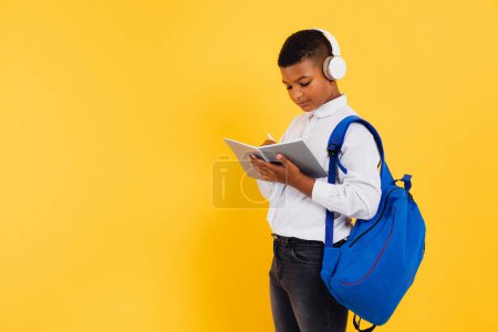 Téléchargez les photos : Heureux écolier mixte portant des écouteurs et sac à dos tenant des livres et des cahiers. Concept de retour à l'école. - en image libre de droit