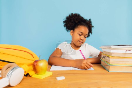 Happy African American schoolgirl doing homework at desk in class on blue background. Back to school concept. Poster 643673972