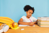Happy African American schoolgirl doing homework at desk in class on blue background. Back to school concept. t-shirt #643673972