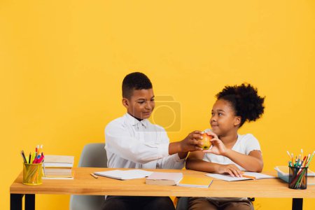 Photo for Happy african american schoolgirl and mixed race schoolboy sitting together at desk and sharing apple on yellow background. Back to school concept. - Royalty Free Image