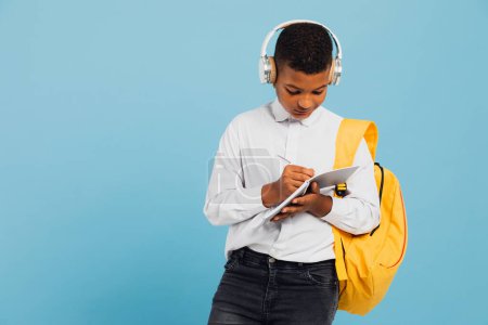 Téléchargez les photos : Heureux écolier mixte portant des écouteurs et sac à dos écrit quelque chose dans un carnet. Concept de retour à l'école. - en image libre de droit