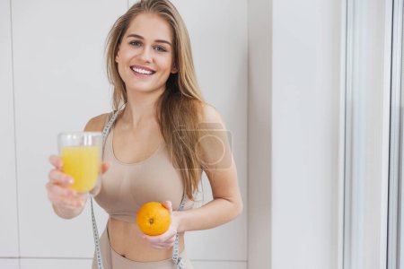 Photo for Happy attractive millennial woman in sportswear with measuring tape holding fresh juice and orange for energy recovery after workout. Copy space - Royalty Free Image