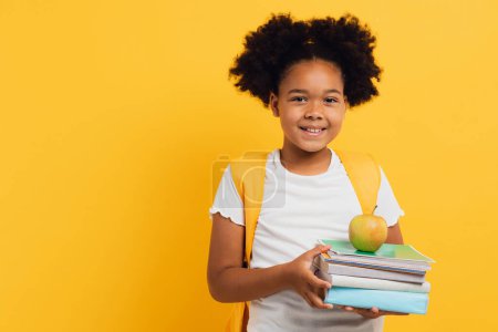 Téléchargez les photos : Heureuse écolière afro-américaine tenant des cahiers et des livres sur fond jaune, espace de copie. - en image libre de droit