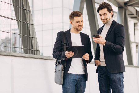 Foto de Dos modernos empresarios felices con teléfono y carpeta de documentos discutiendo algo en el contexto de oficinas urbanas y edificios - Imagen libre de derechos