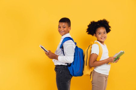 Foto de Feliz colegiala afroamericana y colegial sosteniendo libros uno al lado del otro sobre fondo amarillo. Regreso al concepto escolar. - Imagen libre de derechos