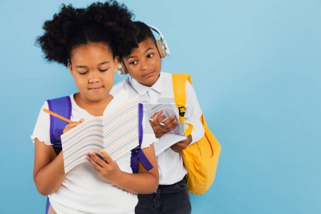 Téléchargez les photos : Un écolier africain distribue ses devoirs à une écolière afro-américaine en classe sur fond bleu, concept de retour à l'école. - en image libre de droit