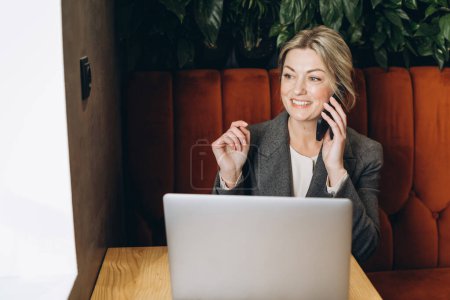 Téléchargez les photos : Femme d'affaires mature administrateur de restaurant assis parlant au téléphone et travaillant sur ordinateur portable - en image libre de droit