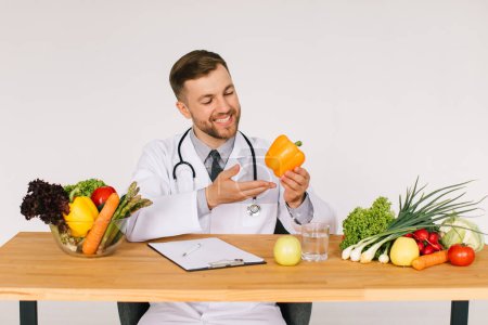 Téléchargez les photos : Heureux médecin nutritionniste assis sur le lieu de travail au bureau parmi les légumes frais et tenant le poivre, concept de plan de régime - en image libre de droit