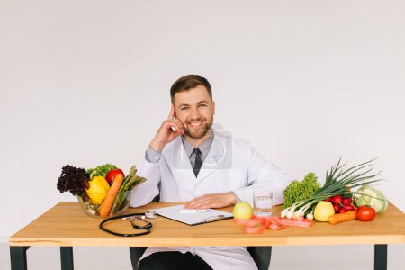 Téléchargez les photos : Heureux médecin nutritionniste assis sur le lieu de travail au bureau parmi les légumes frais, concept de plan de régime - en image libre de droit