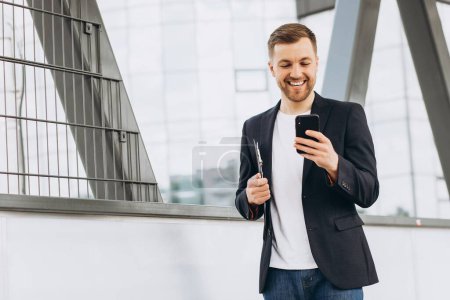 Photo for Portrait of happy modern male businessman in suit walking smiling and reading or writing message on phone on background of urban buildings and offices. - Royalty Free Image