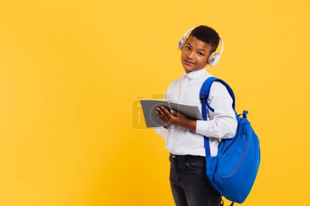 Téléchargez les photos : Heureux écolier mixte portant des écouteurs et sac à dos tenant des livres et des cahiers. Concept de retour à l'école. - en image libre de droit