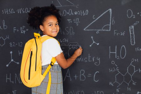 Foto de Feliz colegiala afroamericana resolviendo problemas cerca de la pizarra en la escuela, de vuelta al concepto escolar. - Imagen libre de derechos