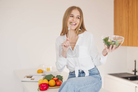 Happy and smiling blonde woman sitting on top of table in kitchen and enjoying homemade salad among cooking ingredients.