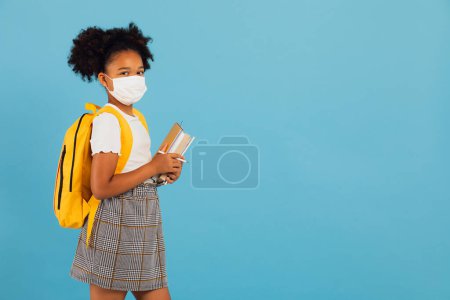 Foto de Colegiala afroamericana en máscara sosteniendo libros sobre fondo azul con espacio para copiar. De vuelta al concepto escolar. COVID-19 - Imagen libre de derechos