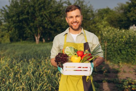 Foto de Hombre agricultor feliz sosteniendo cesta con verduras frescas en el arco iris y el fondo del jardín, concepto de jardinería - Imagen libre de derechos