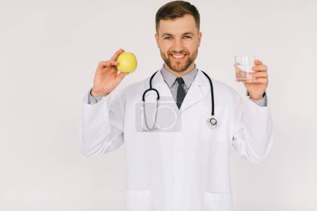 Téléchargez les photos : Le médecin nutritionniste masculin heureux avec stéthoscope souriant et tenant l'eau et la pomme sur fond blanc, concept de plan de régime - en image libre de droit