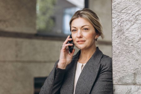 Téléchargez les photos : Portrait d'une belle femme d'affaires mature souriante et parlant au téléphone sur le fond urbain moderne - en image libre de droit