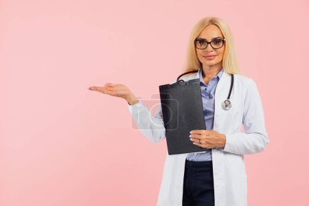 Photo for Mature woman doctor in glasses with stethoscope holding folder and show hand something on copy space the pink background - Royalty Free Image
