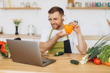 Photo for Happy man in headphones sitting at kitchen table and preparing salad, holding pepper and showing it to laptop. - Royalty Free Image