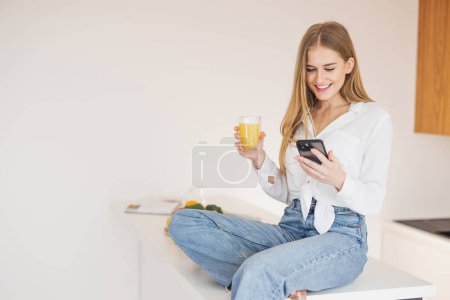 Foto de Mujer rubia feliz y sonriente sentada encima de la mesa de la cocina bebiendo jugo de naranja y hablando por teléfono - Imagen libre de derechos