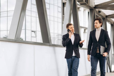 Foto de Dos hombres de negocios modernos con teléfono y carpeta de documentos discutir algo y caminar a través del puente en el fondo de oficinas urbanas y edificios - Imagen libre de derechos