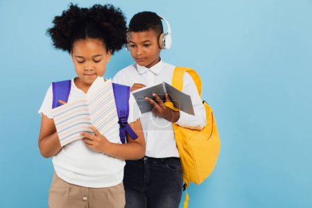 Téléchargez les photos : Un écolier métis distribue ses devoirs à une écolière afro-américaine en classe sur fond bleu, concept de retour à l'école. - en image libre de droit