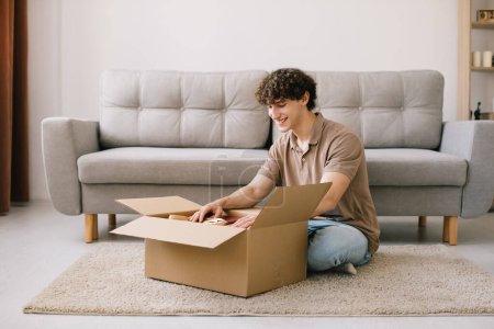 Photo for Happy young smiling curly man opening box with ordered goods gifts, presents at home on couch. Online shopper male customer opening online shop parcel. - Royalty Free Image
