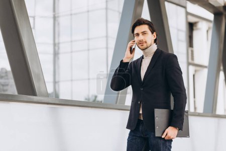 Photo for Portrait of handsome modern male businessman in suit holding folder with documents and talking on the phone against the background of urban buildings and offices. - Royalty Free Image