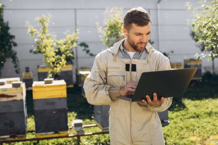 Téléchargez les photos : Homme agronome ou ingénieur en combinaison de protection travaillant sur ordinateur portable à la ferme apicole - en image libre de droit