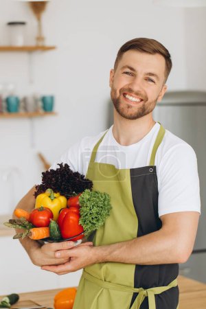 Photo for Portrait of a happy man holding a plate of fresh vegetables on the background of the kitchen at home - Royalty Free Image