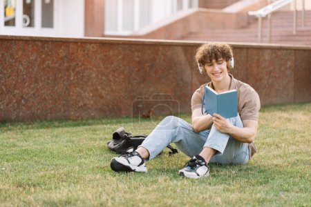 Photo for Attractive male student in headphones studying near the university sitting on the grass, preparing for exams - Royalty Free Image