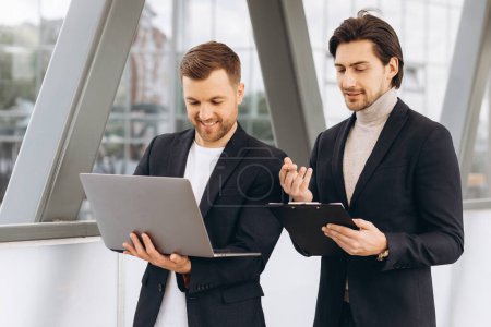 Photo for Two modern happy businessmen with laptop and document folder discussing something against the background of urban offices and buildings - Royalty Free Image
