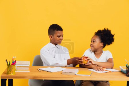 Photo for Happy african american schoolgirl and schoolboy sitting together at desk and sharing apple on yellow background. Back to school concept. - Royalty Free Image