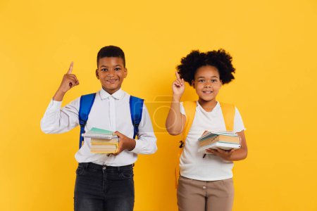 Téléchargez les photos : Heureuse écolière afro-américaine et écolier tenant des livres et montrant quelque chose ensemble sur fond jaune, espace de copie. - en image libre de droit