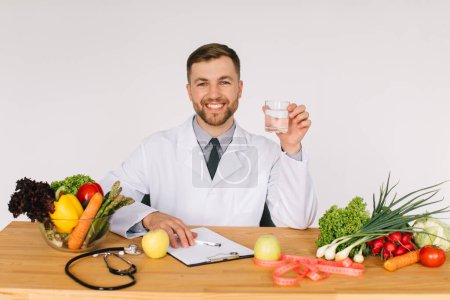 Téléchargez les photos : Heureux médecin nutritionniste assis sur le lieu de travail au bureau parmi les légumes frais tenant un verre d'eau, concept de plan de régime - en image libre de droit