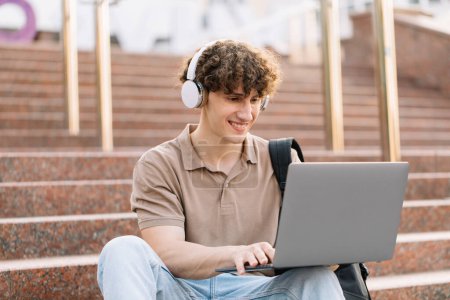 Photo for Concept of e-learning, distance study or remote learning concept, Young happy curly hair school guy, college or university student with backpack and headphones using laptop sitting on steps in university - Royalty Free Image