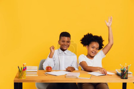 Photo for Happy african american female schoolgirl and schoolboy sitting together at desk and stretching arms up on yellow background. Back to school concept. - Royalty Free Image