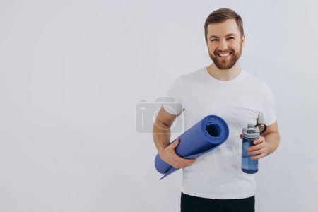 Porträt eines gutaussehenden bärtigen Mannes in Sportbekleidung mit Yogamatte und Flasche Süßwasser auf weißem Hintergrund, Kopierraum