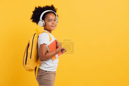Téléchargez les photos : Heureuse écolière afro-américaine dans un casque avec sac à dos tenant des cahiers sur fond jaune, espace de copie. - en image libre de droit