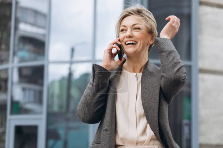 Photo for Portrait of a beautiful mature business woman in suit and gray jacket smiling and talking on a phone on the modern urban and office buildings background - Royalty Free Image