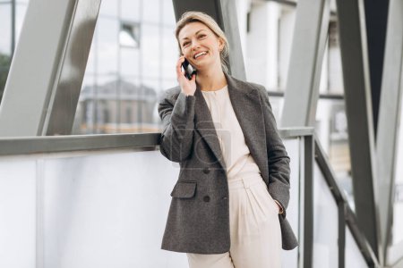 Téléchargez les photos : Portrait d'une belle femme d'affaires mature en costume et veste grise souriante et parlant au téléphone sur le fond urbain moderne - en image libre de droit