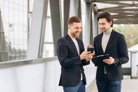 Photo for Two modern businessmen are discussing something and showing each other on a smartphone against the backdrop of urban offices and buildings - Royalty Free Image