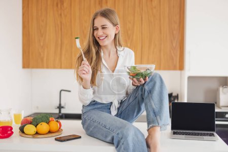 Foto de Mujer rubia feliz y sonriente sentada encima de la mesa de la cocina y comiendo ensalada casera, portátil con espacio para copiar - Imagen libre de derechos