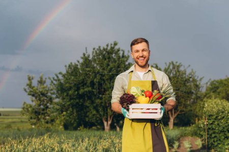 Photo for Happy farmer man holding basket with fresh vegetables on rainbow and garden background, gardening concept - Royalty Free Image
