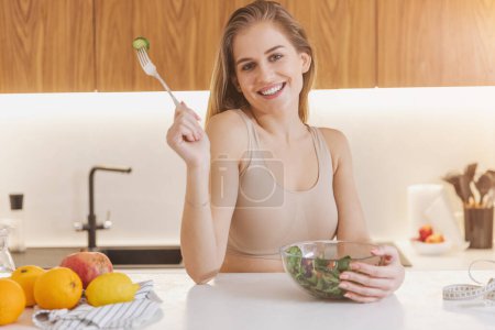 Téléchargez les photos : Heureuse femme millénaire attrayante en vêtements de sport avec ruban à mesurer manger salade de légumes frais pour la récupération d'énergie après l'entraînement dans la cuisine - en image libre de droit