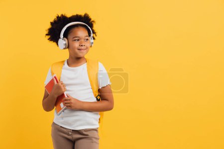 Photo for Happy african american schoolgirl in headphones with backpack holding notebooks on yellow background, copy space. - Royalty Free Image