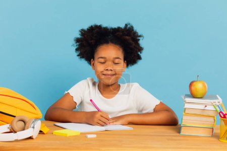 Foto de Feliz colegiala afroamericana soñando con los ojos cerrados y haciendo la tarea en el escritorio sobre fondo azul. Regreso al concepto escolar. - Imagen libre de derechos