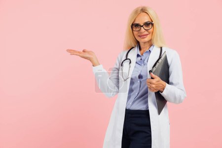 Foto de Mujer madura médico en gafas con estetoscopio celebración de la carpeta y mostrar la mano algo en el espacio de copia el fondo rosa - Imagen libre de derechos