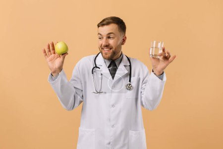 Foto de El médico nutricionista masculino con estetoscopio sonriendo y sosteniendo el agua y la manzana sobre fondo beige, concepto de plan de dieta - Imagen libre de derechos