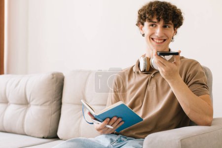 Foto de Joven estudiante masculino de pelo rizado atractivo que estudia sentado en el sofá en casa en la sala de estar, hombre grabando mensaje de voz - Imagen libre de derechos